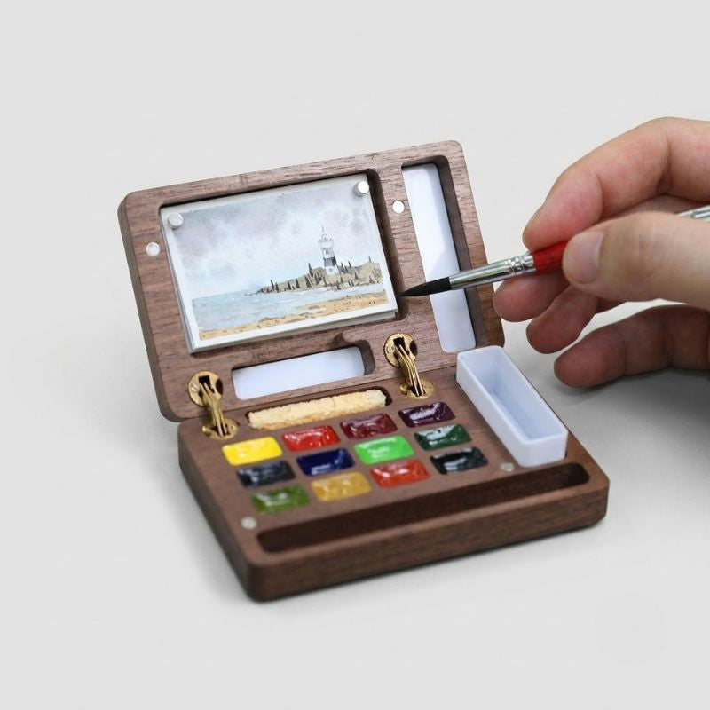 Open ArtMate handmade walnut watercolor palette on a wood rail outdoors, painting a seascape with 12 paint wells