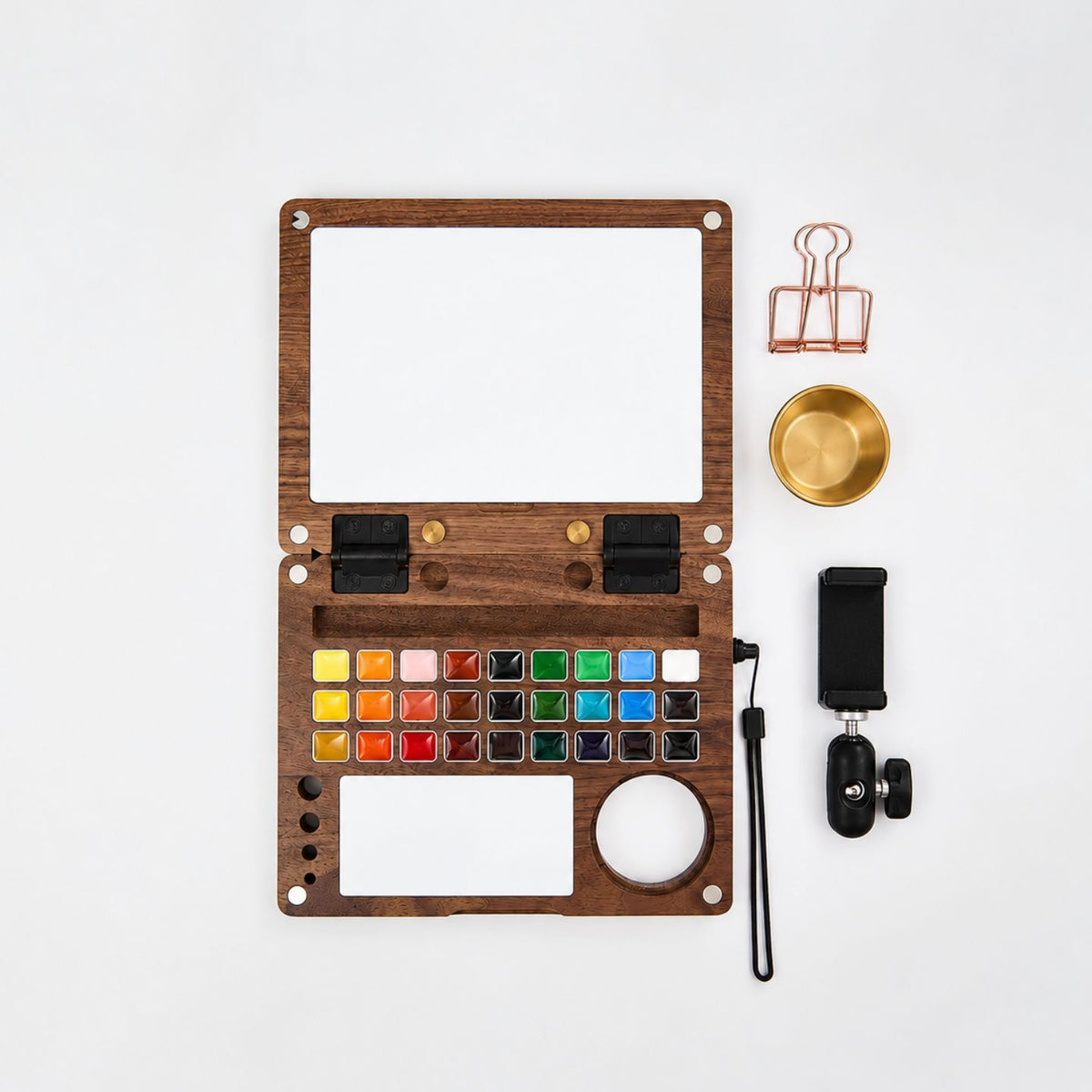 Flat lay of the open walnut Master Edition kit — 27-pan palette, mixing tile and water-pot well — beside a brass water pot, rose-gold binder clip, wrist strap and phone-holder clamp
