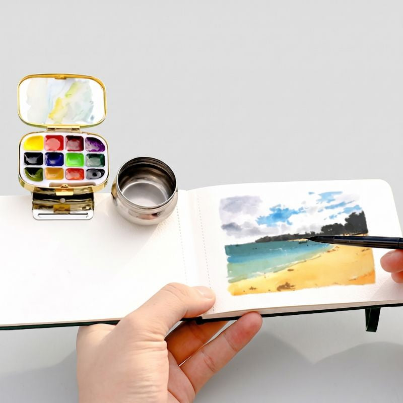 Open ArtMate Gold Edition watercolor box with 12 pans and a metal water cup beside a beach painting in a sketchbook