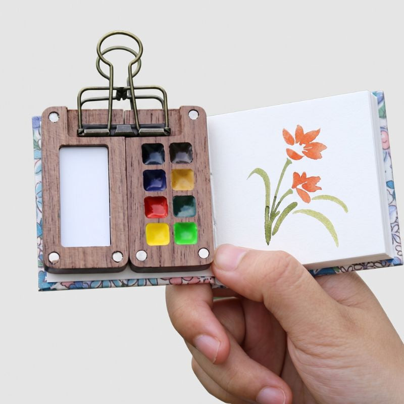 Hand holding the open ArtMate Mini Adventure Artist Kit, a walnut 8-color palette with a floral sketchbook and a painted flower