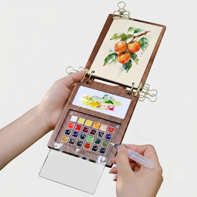 Hands holding the open ArtMate walnut art journal outdoors, painting persimmons with the 24-color palette